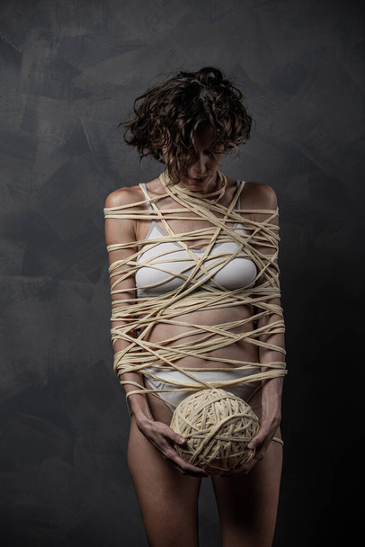 Beautiful woman wrapped in a rope with a ball of thread over grey wall - Φωτογραφία, εικόνα