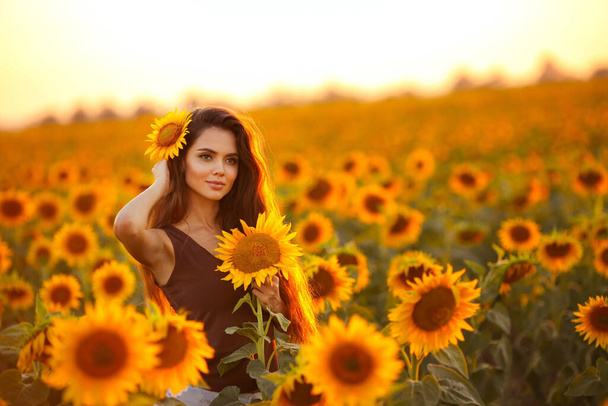 Beautiful young girl enjoying nature on the field of sunflowers at sunset. Summertime. Attractive brunette woman with long healthy hair. - Foto, Imagem