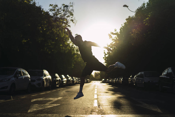 A young sporty guy jumping in the middle of an asphalt road at sunset - Фото, изображение