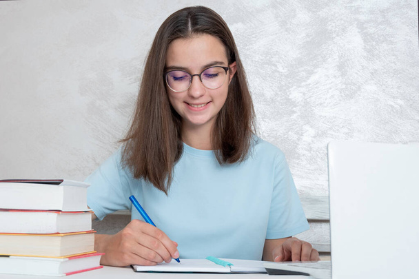 A smiling teenager student sitting at a table with books is writing an assignment in a notebook. - Photo, Image
