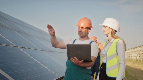 Technik a investor procházející solární buňkou Farma přes pole solárních panelů kontrolující panely v solární energetice. Muž drží notebook v rukou stojící na poli se solárními panely. - Fotografie, Obrázek