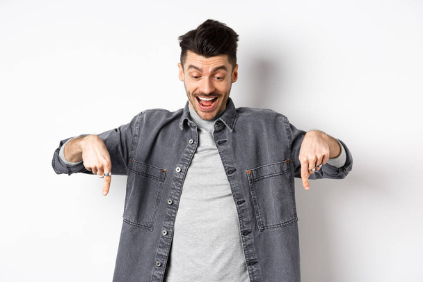 Aufgeregter europäischer Mann, der in Jeansjacke auf weißem Hintergrund steht und mit den Fingern nach unten zeigt, glücklich auf die Werbung blickt - Foto, Bild
