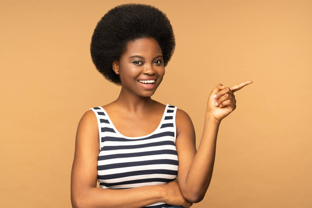 Happy african woman point finger left with excited cheerful smile. Young black girl with afro hairstyle present show new product or sale advertising space isolated over beige studio wall background - 写真・画像