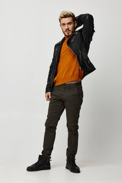 homem sexy em uma jaqueta de couro segura a mão atrás de sua cabeça e luz fundo laranja calças de camisola - Foto, Imagem