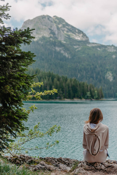 Young woman on shore of mountain lake. The back of girl against background of mountains. Young girl sitting near by beautiful Black Lake in Montenegro. Original wallpaper from summer morning - Foto, afbeelding