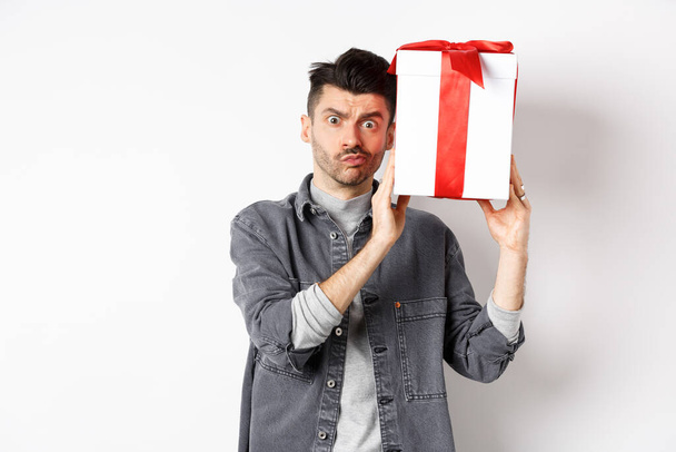 Intrigued young man looking confused at camera, trying to guess what inside gift box from lover, shaking present and stare puzzled, standing on white background. Valentines day concept - Photo, Image