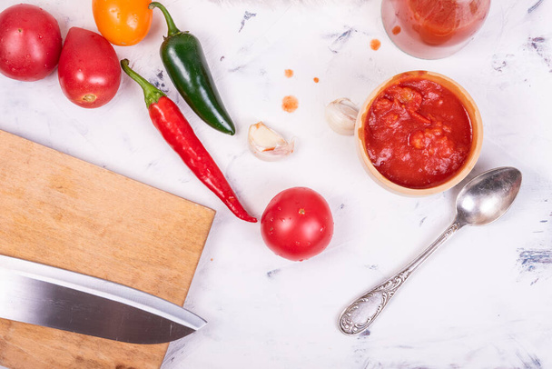 Ainesosien asettelu salsakastikkeen valmistukseen - hienonnettuja tomaatteja kastikkeessa, tomaatteja, tulisia paprikoita ja valkosipulia, leikkuulauta veitsellä - Valokuva, kuva