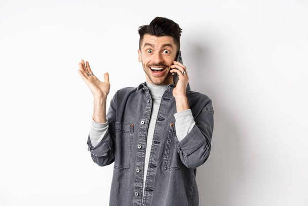 Nadšený muž mluví na mobilu a slyší dobré zprávy, raduje se a zvedá ruku se šťastným úsměvem, stojí na bílém pozadí - Fotografie, Obrázek