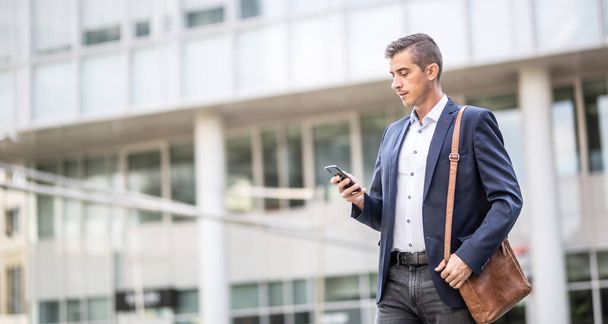 Homem em traje de negócios casuais verifica seu telefone andando ao ar livre em um ambiente de edifícios de escritório. - Foto, Imagem