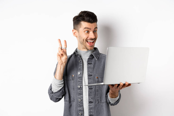 Funny young man video chat on laptop, showing v-sign and smiling at computer camera, standing against white background - 写真・画像