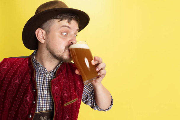 Close-up young man dressed in traditional Bavarian costume drinking cold foamed beer, lager, ale isolated over yellow background. National cuisine, holidays, traditions - Fotografie, Obrázek