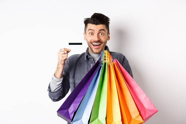 Cara feliz shopaholic segurando sacos de compras coloridos e mostrando cartão de crédito de plástico, sorrindo animado, de pé fundo branco - Foto, Imagem