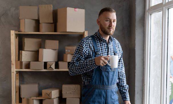 young Caucasian small business owner stands near a rack with boxes and holds a cup of coffee. online sales, e-commerce and small and medium business concept - Photo, Image
