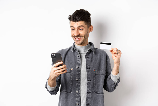 Concept d'achat en ligne. Un gars souriant payant sur son téléphone portable, montrant une carte de crédit en plastique, achetant sur Internet, fond blanc - Photo, image