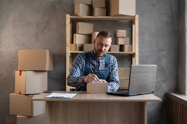 Portrait of a small business startup owner working at home with laptop. Online sale. A Caucasian man sits at a table against the backdrop of a rack with boxes and writes the delivery address with a pen. - Photo, Image