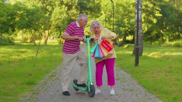 Anciano elegante pareja abuela, abuelo después de comprar con bolsas usando scooter para montar a caballo - Metraje, vídeo