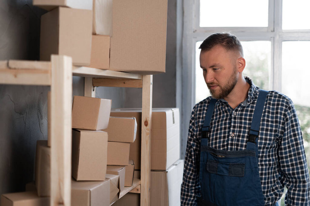 Young man dressed in blue overalls near cardboard boxes in a warehouse. Work in a logistics company. - Photo, Image