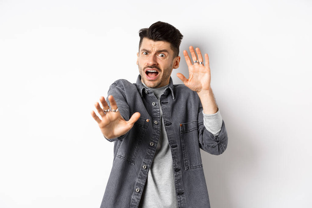 Scared timid guy screaming and raising hands in pleading pose, begging to stop, defending from assault, standing on white background - Фото, изображение