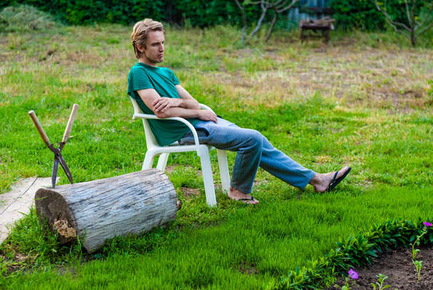 Δούλεψε στον κήπο. Χλόουν. Ένας άντρας κάθεται σε μια καρέκλα στο γρασίδι δίπλα σε ένα παρτέρι. Ψαλίδι κηπουρικής. - Φωτογραφία, εικόνα