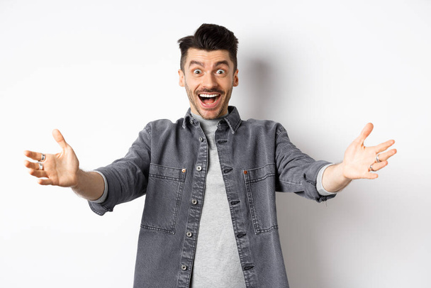 Excited young man stretch out hands for hug, cuddling you, welcoming guest and smiling, inviting people, standing on white background - Photo, image
