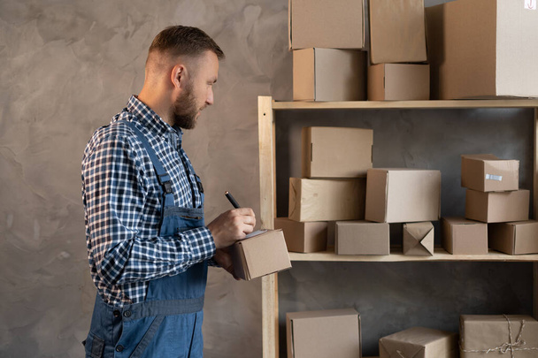 Caucasian male worker dressed in a blue overalls stands near a shelf with boxes makes a pen marks on the box. Copy space. I work in the Logistics and Distribution Center. - Photo, Image