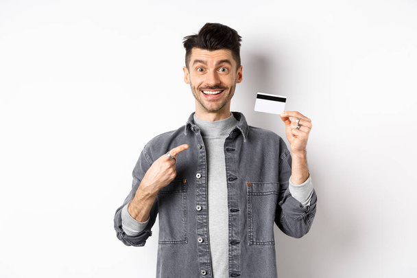 Opgewonden knappe man met snor wijzende vinger naar plastic creditcard, glimlachend blij, aanbevelen goede deal, staan op witte achtergrond - Foto, afbeelding
