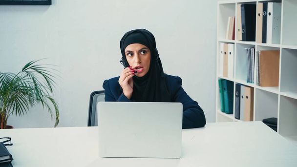 Muslim woman in headset using microphone near laptop in call center  - 写真・画像