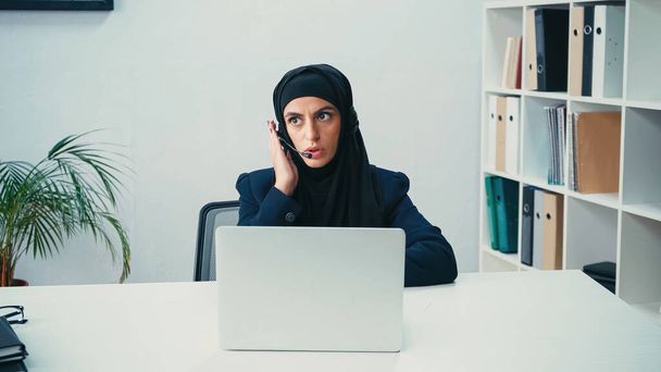 Muslimische Frau in Kopfhörer mit Mikrofon und Gespräch in der Nähe von Laptop in Call Center  - Foto, Bild
