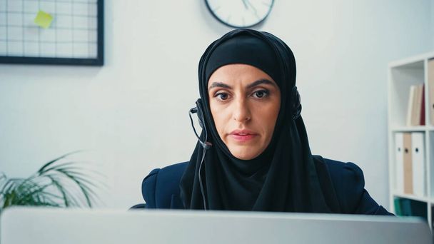 Mujer musulmana en hijab usando auriculares y mirando borrosa computadora portátil en el centro de llamadas  - Foto, Imagen