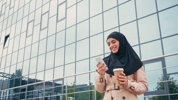 low angle view of happy muslim woman in hijab using smartphone and holding coffee to go near building  - Photo, Image