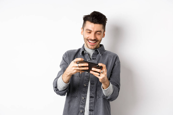 Красивий молодий чоловік грає в мобільну відеогру і посміхається на екрані, стоячи на білому тлі
 - Фото, зображення
