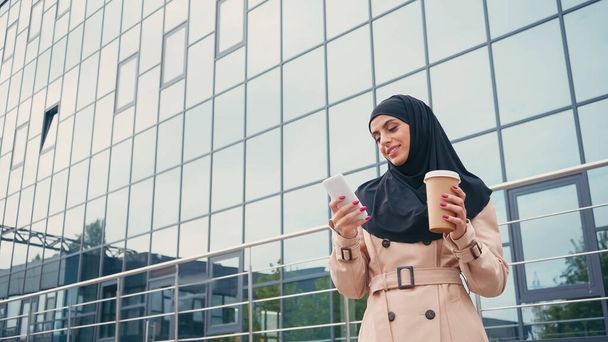 веселая молодая мусульманка в хиджабе с помощью смартфона и держа кофе, чтобы подойти к зданию  - Фото, изображение