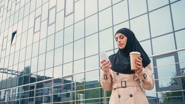 jovem mulher muçulmana no hijab usando smartphone e segurando café para ir perto do edifício  - Foto, Imagem