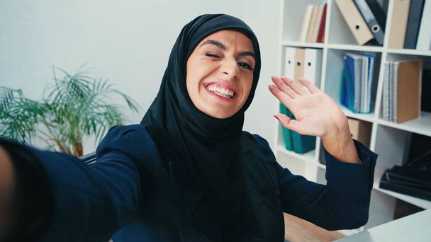 Smiling muslim businesswoman waving hand while taking selfie  - Photo, Image