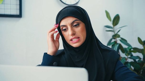 смущенная мусульманка в хиджабе смотрит на размытый ноутбук - Фото, изображение