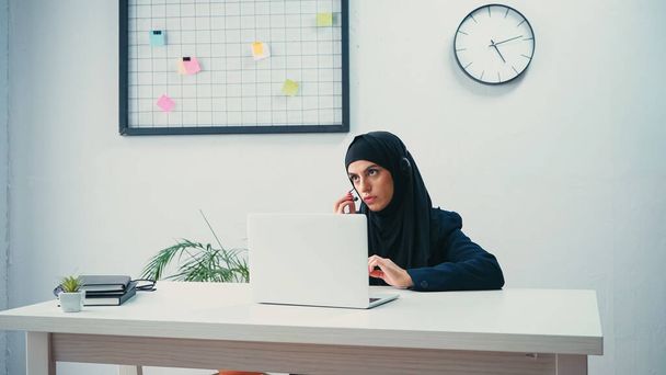 Mujer musulmana en hijab y auriculares que ajustan el micrófono cerca de la computadora portátil en el centro de llamadas  - Foto, Imagen
