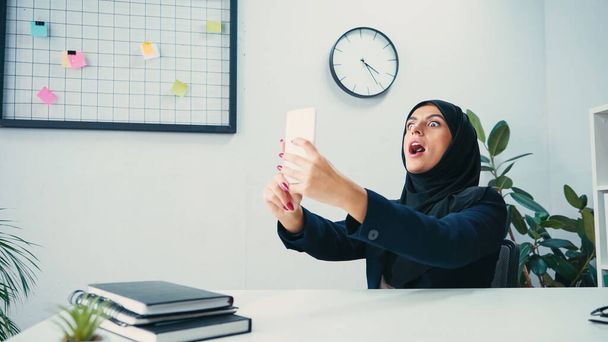 Donna d'affari musulmana a bocca aperta che si fa selfie sullo smartphone in ufficio  - Foto, immagini