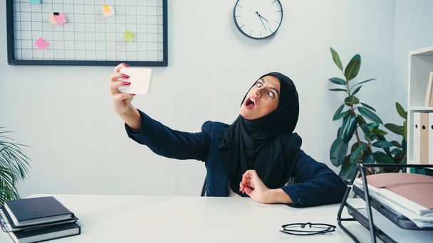 Mulher de negócios muçulmana sorrindo enquanto toma selfie no smartphone no escritório  - Foto, Imagem