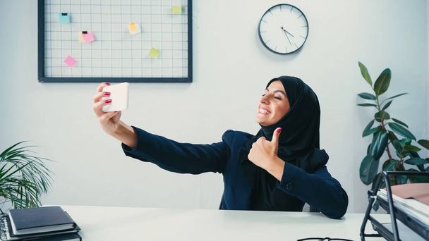 feliz musulmana mujer de negocios mostrando el pulgar hacia arriba mientras toma selfie en el teléfono inteligente en la oficina  - Foto, Imagen