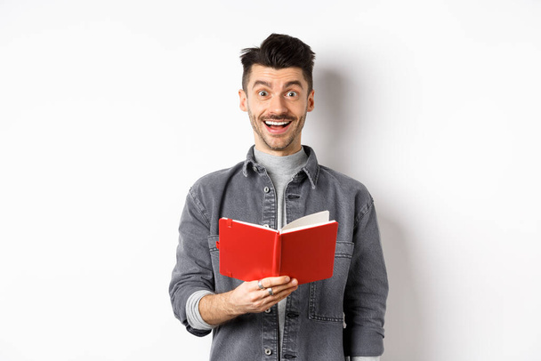Хобби и досуг. Счастливый молодой человек читает планировщик, ведет дневник или красный дневник и улыбается, делает заметки, стоя на белом фоне - Фото, изображение