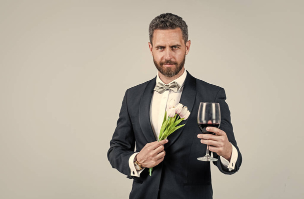 handsome tuxedo man in bow tie with flower bouquet greeting with glass of red wine, happy valentines day, copy space - Fotó, kép