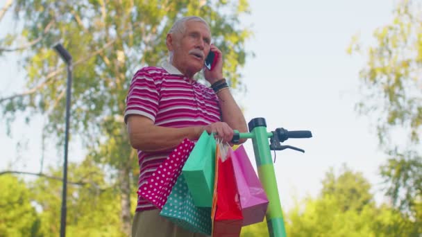 Anciano abuelo hombre en scooter eléctrico con coloridas bolsas de compras hablando en el teléfono móvil - Metraje, vídeo