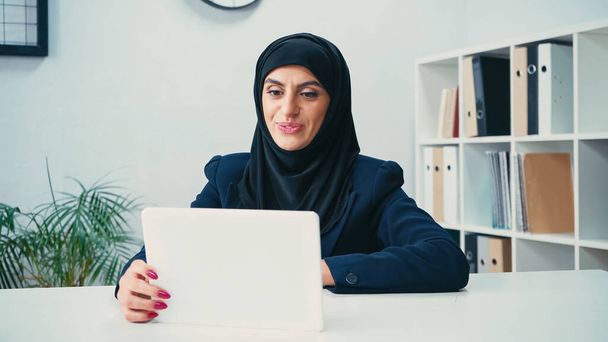 χαμογελαστή μουσουλμάνα γυναίκα σε hijab κοιτάζοντας ψηφιακή ταμπλέτα στο γραφείο  - Φωτογραφία, εικόνα