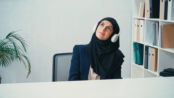 verträumte arabische Geschäftsfrau im Hidschab hört im Büro Musik über Kopfhörer  - Foto, Bild