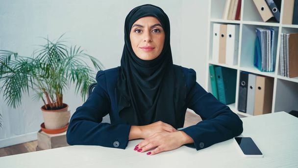 Νεαρή μουσουλμάνα γυναίκα κοντά smartphone με λευκή οθόνη στο γραφείο - Φωτογραφία, εικόνα