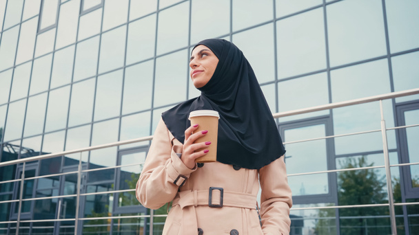 vista de ángulo bajo de la mujer musulmana en hijab y gabardina sosteniendo café para ir cerca de la construcción exterior  - Foto, Imagen