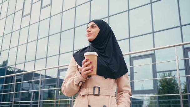 Türbanlı ve trençkotlu genç Müslüman kadının binaya yaklaşmak için elinde kahve tutarken düşük açılı görüntüsü  - Fotoğraf, Görsel