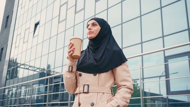 низкий угол зрения мусульманской женщины в хиджабе и плащ держа бумажную чашку возле здания снаружи  - Фото, изображение