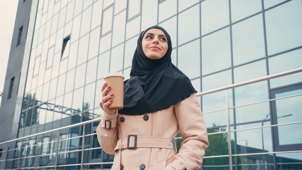 низкий угол зрения веселой мусульманской женщины в хиджабе и траншеи пальто держа бумажную чашку возле здания снаружи  - Фото, изображение