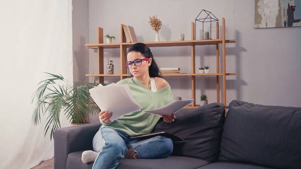 Concentrated student in eyeglasses looking at documents on couch in living room  - Foto, Imagem